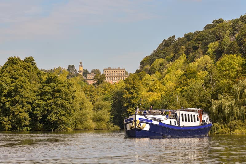 Hotel barge Magna Carta cruising on the River Thames behind Cliveden House